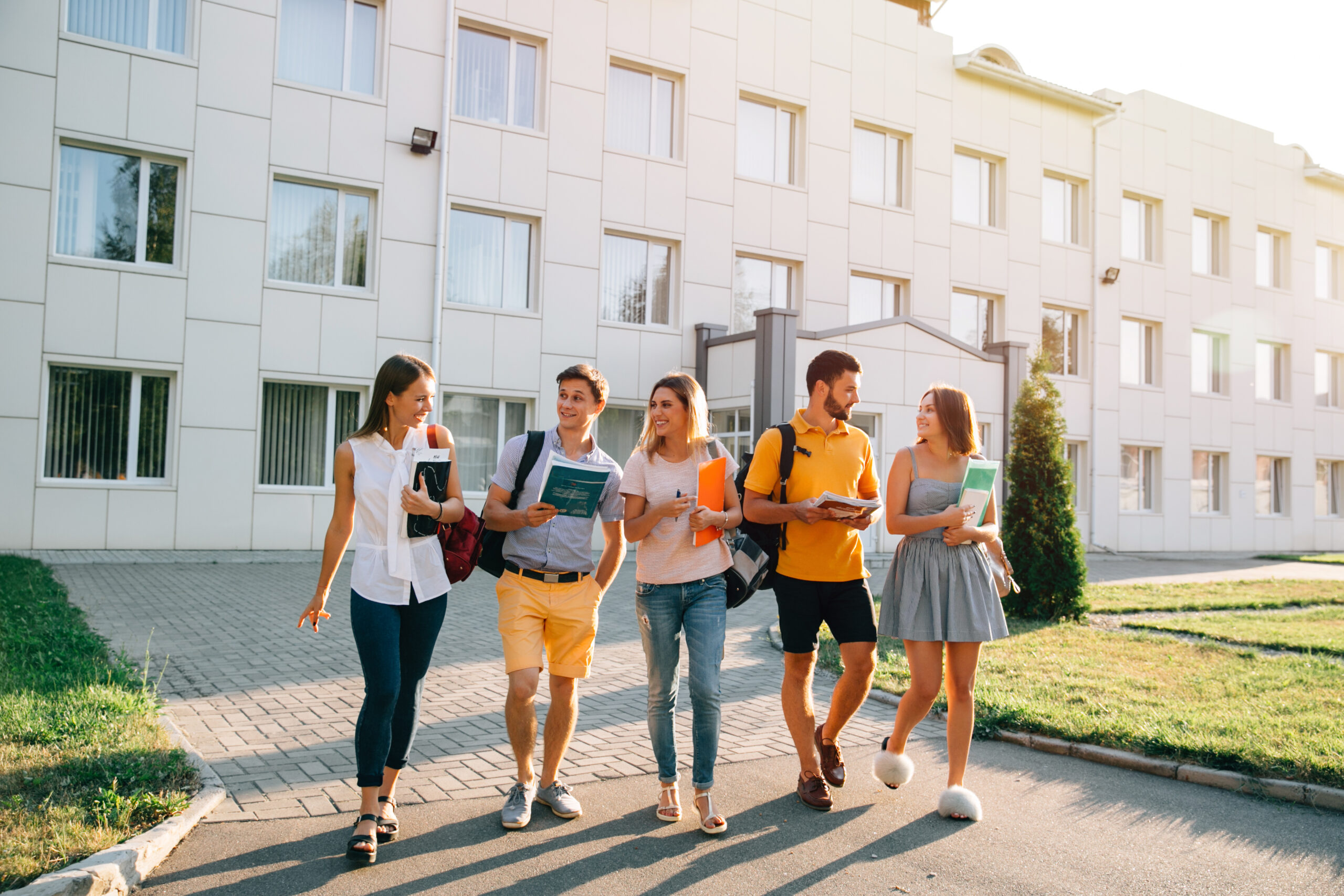 Free time of a students, bachelor`s campus life rhythm. Five friendly students are walking after they passed test outside the college building and discuss the project, smiling, enjoying, carefree