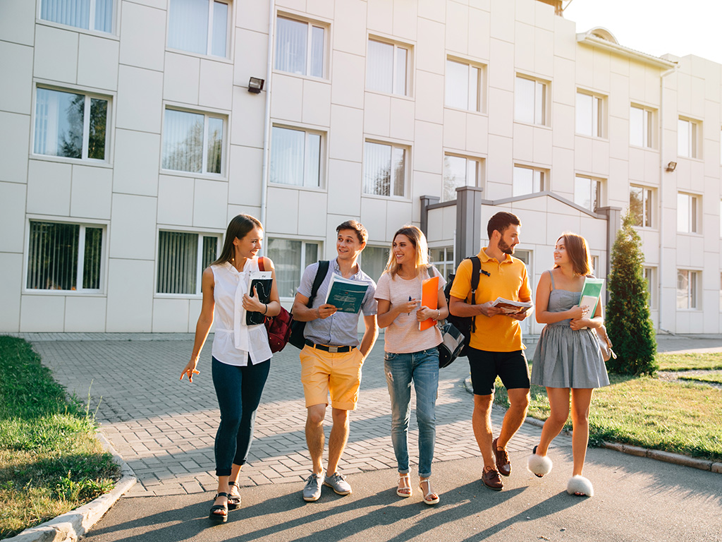 Free time of a students, bachelor`s campus life rhythm. Five friendly students are walking after they passed test outside the college building and discuss the project, smiling, enjoying, carefree